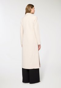 Lungo cappotto crema con colletto a revers, texture liscia e silhouette dritta, abbinato a pantaloni neri ampi.