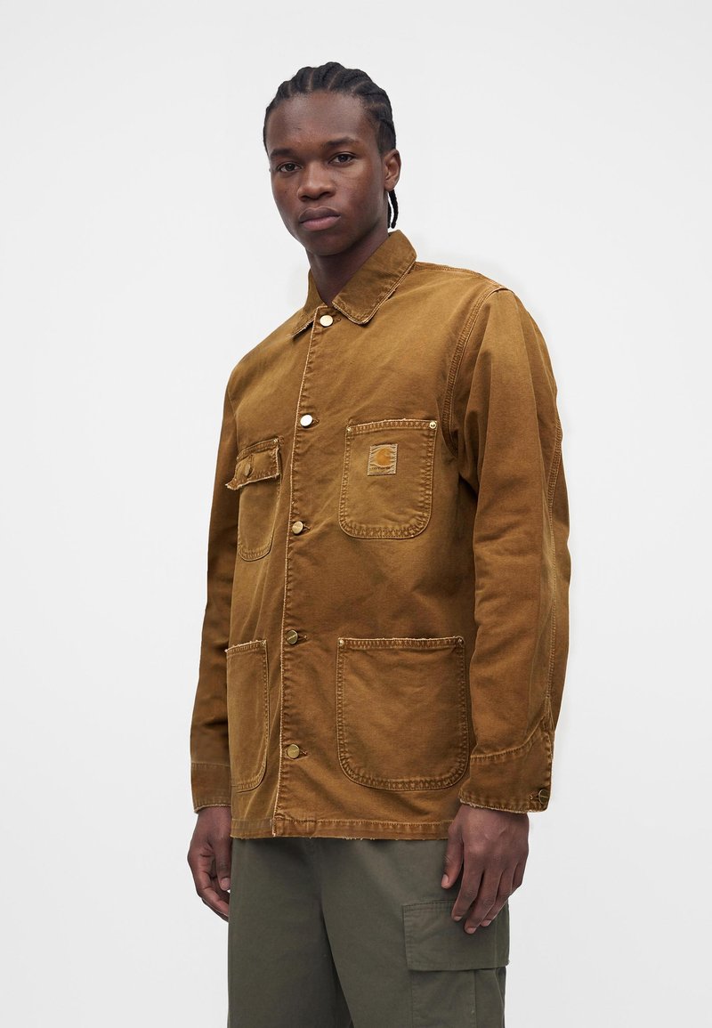 Giovane uomo con capelli intrecciati che indossa una giacca marrone con bottoni e diverse tasche e pantaloni cargo verde oliva.
