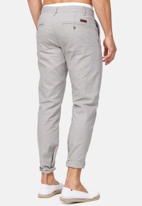 Pantalon en lin gris clair avec un design fuselé, revers roulés, deux poches à l'arrière, et un accent en cuir marron. Porté avec des chaussures blanches plates.
