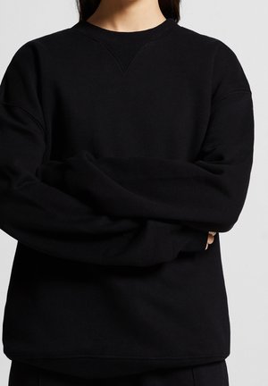 Person trägt ein locker sitzendes schwarzes Sweatshirt mit langen Ärmeln, geripptem Halsausschnitt und Saum, die Arme vor der Brust verschränkt.