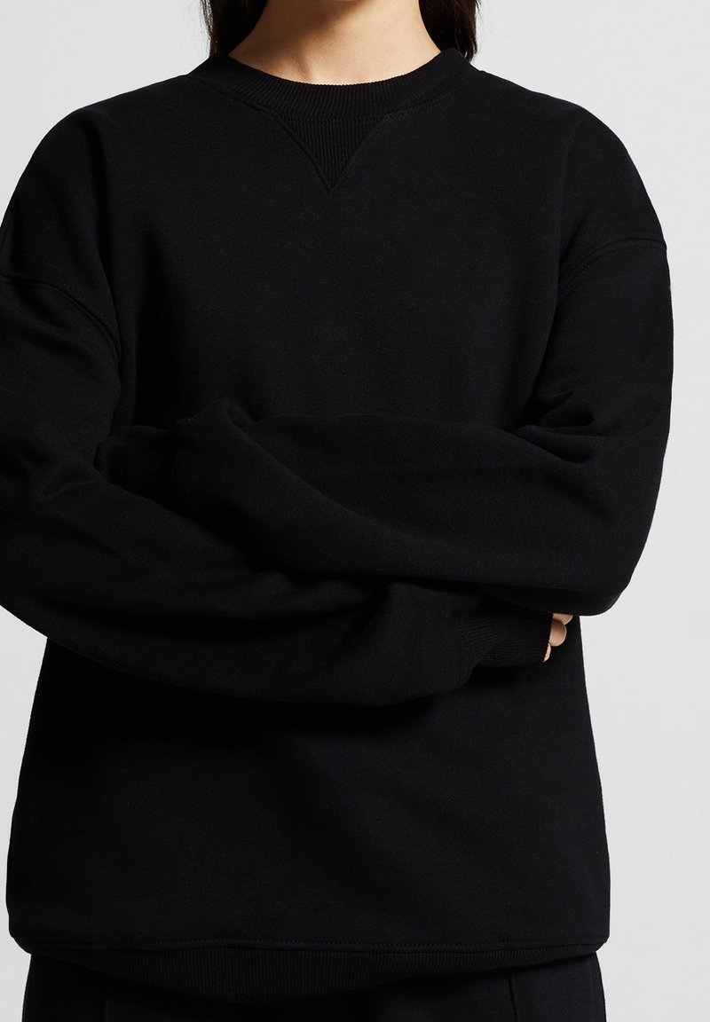 Personne portant un sweat-shirt noir ample à manches longues avec encolure et ourlet côtelés, les bras croisés devant.