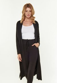 Langer schwarzer Cardigan mit lockerer Passform, offenem Vorderteil, seitlichen Taschen und weicher Textur, getragen über einem weißen Oberteil und schwarzen Hosen.