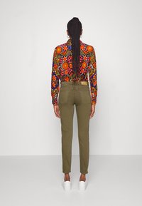 Blusa floral de manga larga con patrón multicolor; pantalones ajustados verde oliva; zapatillas blancas; cabello trenzado; fondo gris liso.