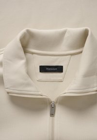 Krémovo sfarbená bunda na zips s mäkkou textúrou, so zloženým golierom a čiernou štítkom s nápisom "Matinique" vo vnútri oblasti krku.