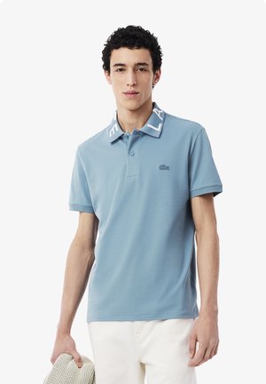 Vaaleansininen polo-paita, jossa on valkoinen kaulus ja "LACOSTE"-teksti. Paidassa on pieni Lacoste-logo rinnassa ja se on sileää kangasta.