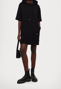 Czarna sukienka z kapturem, krótkimi rękawami, kieszenią kangurką, regulowanym sznurkiem przy kapturze oraz detalem z logo. Noszona z czarnymi botkami za kostkę i czarną torebką.