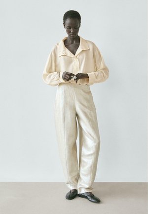 Donna con indosso una camicetta beige ampia e pantaloni larghi lucidi, che si sistema un anello, in piedi davanti a uno sfondo grigio chiaro uniforme.