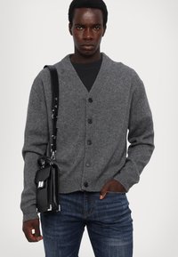 Grau gestrickter Cardigan mit V-Ausschnitt, Knöpfen vorne und gerippten Bündchen. Kombiniert mit dunklen Jeans und einer schwarzen Schultertasche mit metallischen Akzenten.