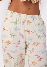 Lichte broek met een elastische tailleband, met een roomkleurige achtergrond en kleurrijke dinosaurusafbeeldingen in roze, blauw, groen en oranje.