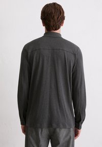 Mężczyzna z krótkimi brązowymi włosami, ubrany w ciemnoszarą koszulę z długim rękawem i szare spodnie, stojący twarzą do gładkiej białej ściany.