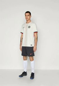 Muž stojaci v bielom futbalovom drese s červenými a zelenými detailmi, čiernych šortkách, bielych ponožkách a čiernych kopačkách na jednoduchom pozadí.