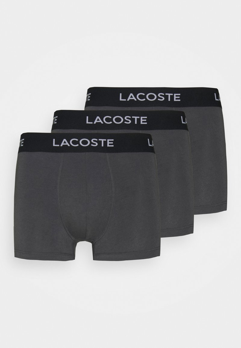 Trois paires de shorts boxers en coton gris avec une taille noire portant le logo "Lacoste" en blanc. Texture lisse, design ajusté.
