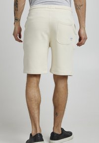 Cremfarbene Sweatshorts aus weichem Material, ausgestattet mit Seitentaschen, einer Gesäßtasche und einem umgeschlagenen Saum. Getragen mit schwarzen Sneakers.