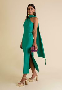 Un vestido de noche verde satinado con cuello halter, abertura lateral y una capa fluida. Acompañado de sandalias de plataforma doradas y un bolso de mano texturizado púrpura.