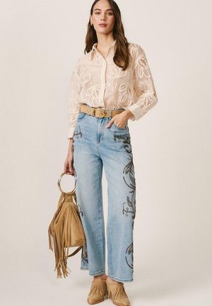 Mujer con blusa transparente de estampado de hojas, jeans bordados de pierna ancha, cinturón marrón, mocasines con flecos, sosteniendo un bolso marrón con flecos y asas de aro.