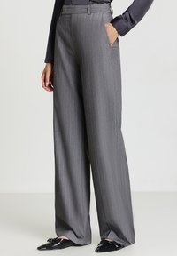 Donna che indossa pantaloni gray a righe verticali a vita alta e gamba larga, con mocassini neri e un top scuro a maniche lunghe, una mano in tasca.
