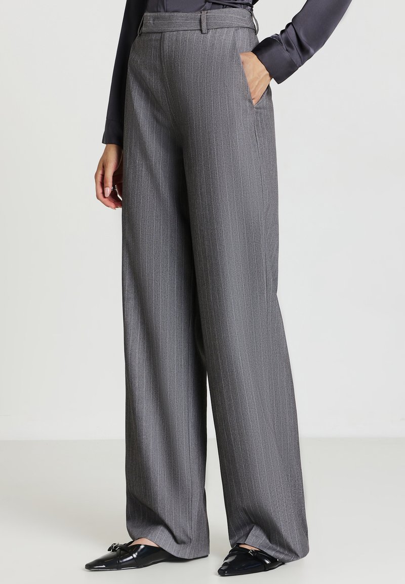 Donna che indossa pantaloni gray a righe verticali a vita alta e gamba larga, con mocassini neri e un top scuro a maniche lunghe, una mano in tasca.