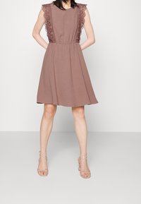 Ruskea hihaton mekko, jossa on teksturoitu kangas, ja pitsikoristeet olkapäillä sekä elastinen vyötärö. Yhdistetty kirkkaisiin hihnakenkiin.