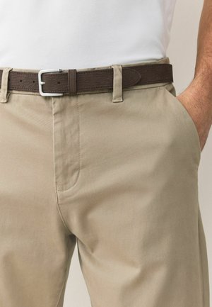 Mężczyzna w beżowych spodniach z ciemnobrązowym skórzanym paskiem i białą koszulą, z ręką częściowo włożoną do prawej kieszeni.