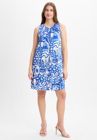 Vrouw in een mouwloze, knie-lange jurk met blauw-witte abstracte patronen, gecombineerd met naakte hoge hakken met enkelband.