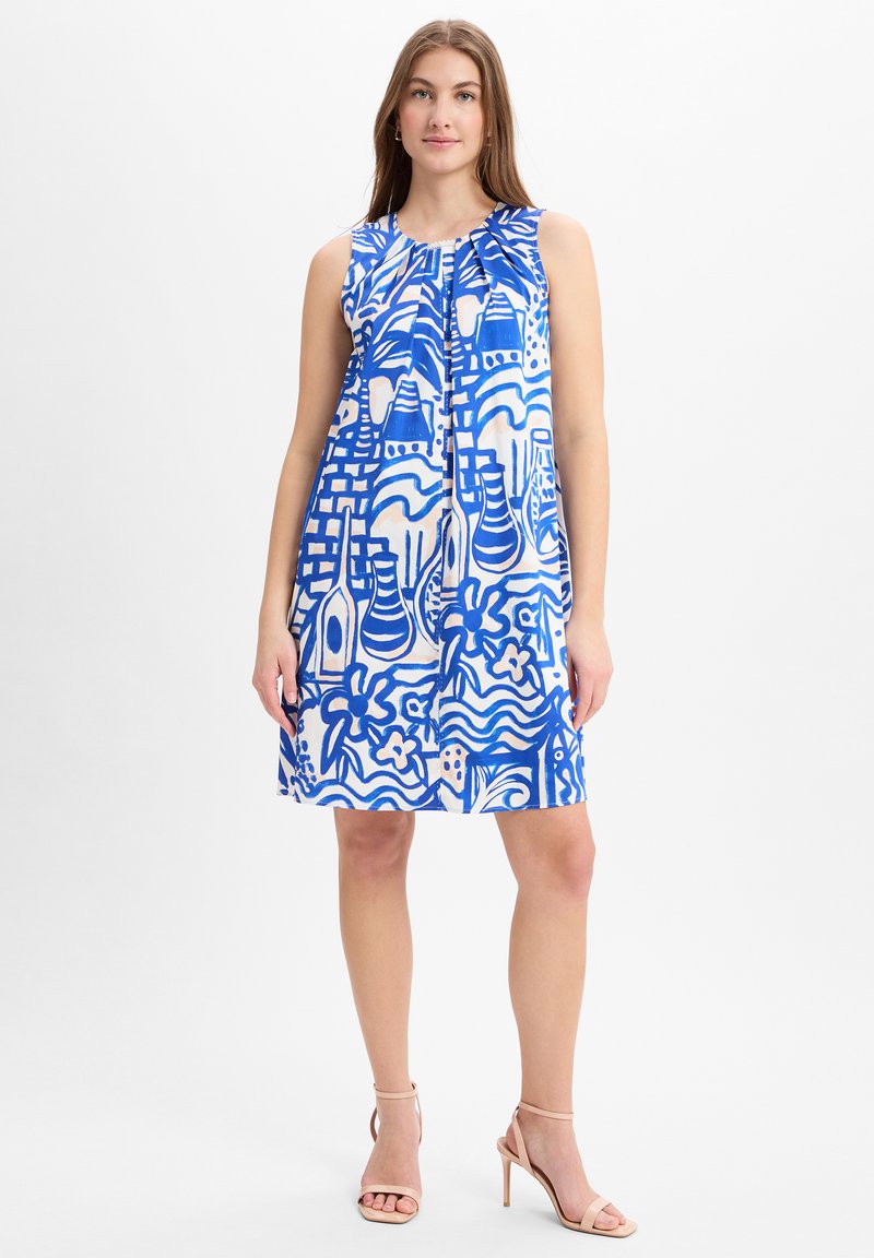 Vrouw in een mouwloze, knie-lange jurk met blauw-witte abstracte patronen, gecombineerd met naakte hoge hakken met enkelband.