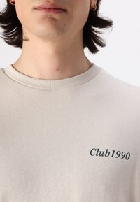 Šviesiai smėlinis džemperis su banguota apykakle, priekyje su tamsiai žaliais užrašais "Club1990", lygus audinio paviršius.