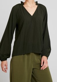 Blusa verde scuro, strutturata, con scollo a V, realizzata in un tessuto leggero, con maniche lunghe arricciate ai polsini, caratterizzata da un delicato motivo crepè.