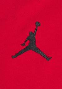 Tessuto rosso con il logo Jumpman ricamato in nero, che ritrae una figura in una posa in aria che tiene un pallone da basket. Texture liscia.
