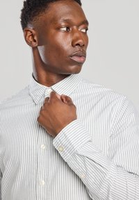 Camisa de botones a rayas en gris claro y blanco, con cuello, que presenta una textura de tela suave y botones con apariencia de perla en la parte frontal y los puños.