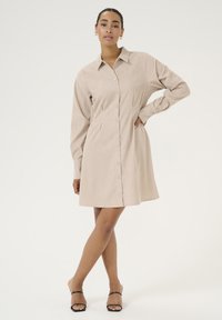 Robe chemise beige avec col, manches longues et fermeture à boutons. Tissu texturé avec poches latérales, tombant au-dessus du genou. Sandales noires.