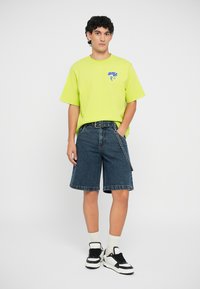 Neon green oversized T-shirt with blue graphic, paired with dark denim shorts and black sneakers with white accents and white socks.