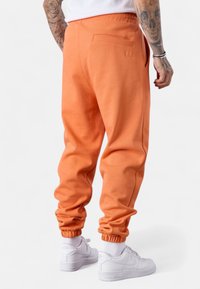 Orange Sweatpants mit schmalem Schnitt, elastischen Bündchen und einer einzelnen Gesäßtasche, kombiniert mit weißen Sneakers. Glatte Baumwolltextur.