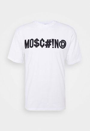 Henkilö nojaa kivikaiteeseen, joka pukeutunut valkoiseen graafiseen Moschino-T-paitaan, mustiin shortseihin, mustiin sukkiin ja mustiin nahkakenkiin, joissa on metallirenkaat.