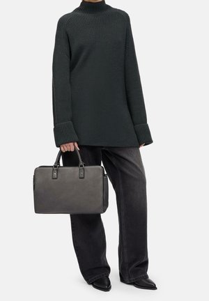 Persona con un suéter de punto verde oscuro y jeans negros de pierna ancha, sosteniendo un maletín de cuero gris por las asas.