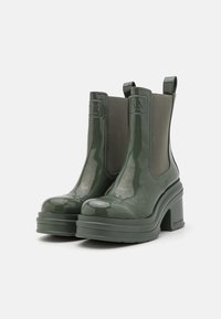 Botas de tobillo de charol verde con gruesos tacones gruesos, paneles laterales elásticos y acentos de logo en el talón y la parte superior. Acabado suave y brillante.
