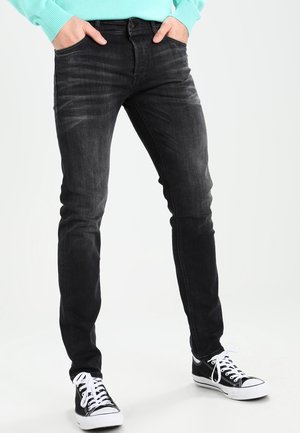Person trägt eine eng anliegende schwarze Jeans, hellblauen Pullover und schwarz-weiße Sneakers, steht mit den Händen in den Taschen vor weißem Hintergrund.