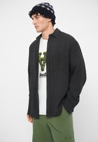 Mørk grå teksturert skjorte med knapper og to frontlommer, båret over en hvit grafisk t-skjorte og kombinert med olivengrønne cargobukser.