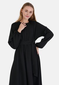 Schwarzes Kleid aus weichem Stoff, mit langen Ärmeln, tailliertem Bund und einem gebundenen Gürtel sowie einem fließenden Rock. Glatte Textur mit minimalistischen Design.