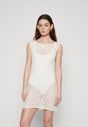 Vit spetsklänning med geometriskt mönster, ärmlös design, rundad halsringning och en något draperad nederkant, med texturerad tyg över hela.