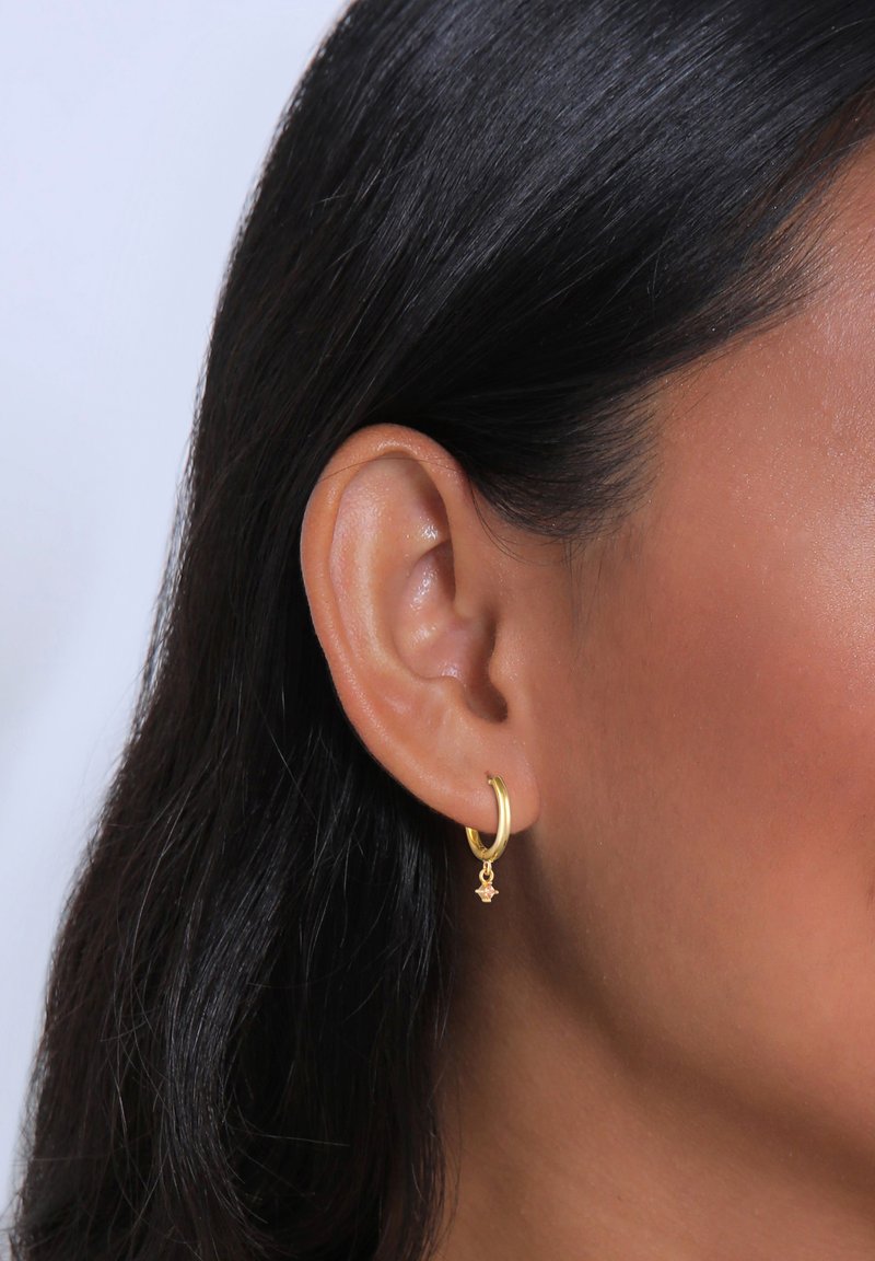 Pendiente de aro dorado con un pequeño charm en forma de estrella colgando de la parte inferior. Superficie lisa, forma circular y acabado pulido.