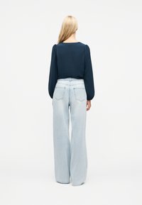 Γυναίκα με μακριά ξανθά μαλλιά, που φορά μπλούζα με μακριά μανίκια σκούρου μπλε και ανοιχτόχρωμα φαρδιά τζιν παντελόνια, γυρισμένη με την πλάτη προς τα εμπρός σε λευκό φόντο.