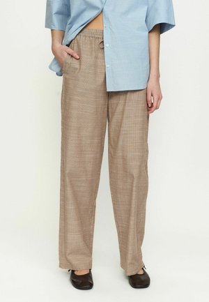 Kobieta ubrana w luźne beżowe spodnie w kratę z elastyczną talią i czarne płaskie buty, w zestawieniu z jasnoniebieską koszulą na pół rozpiętą.