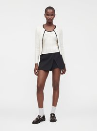 Femme portant un cardigan blanc et un haut avec une bordure noire, un short noir, des chaussettes blanches et des mocassins marron foncé, debout devant un fond blanc.