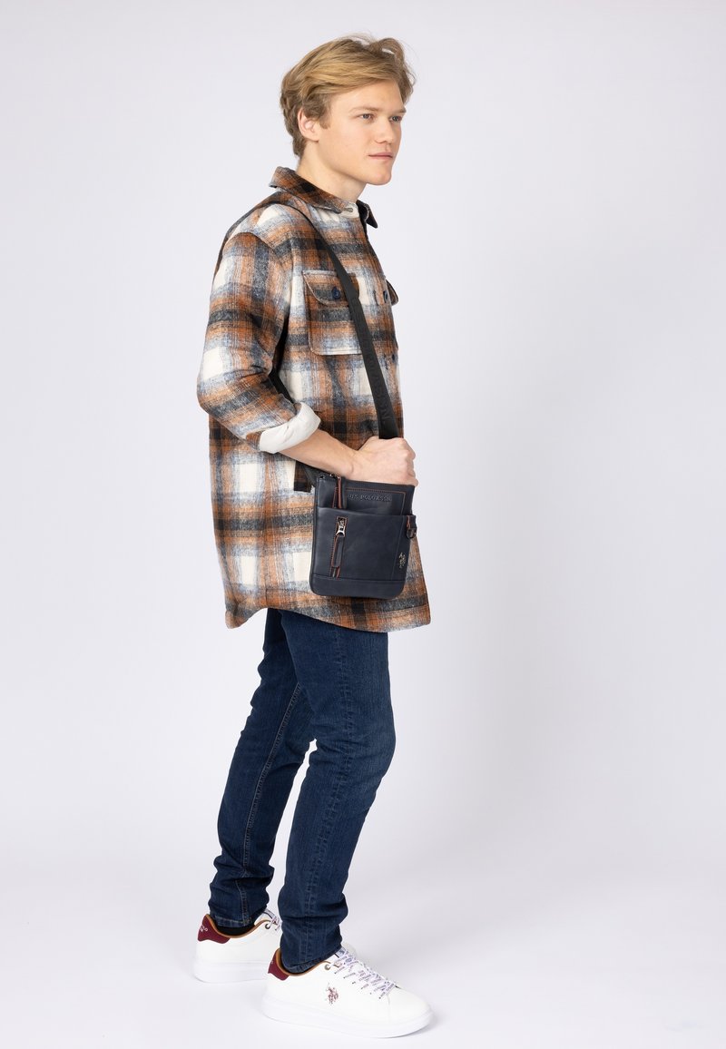 Borsa a tracolla in pelle scura con cerniera, tasca frontale e tracolla lunga. Il modello indossa una camicia a quadri, jeans scuri e sneakers bianche.