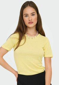 Młoda kobieta z długimi brązowymi włosami, ubrana w żółtą bluzkę z krótkim rękawem, czarną spódnicę i naszyjnik z pereł z muszlami i gwiazdkami.