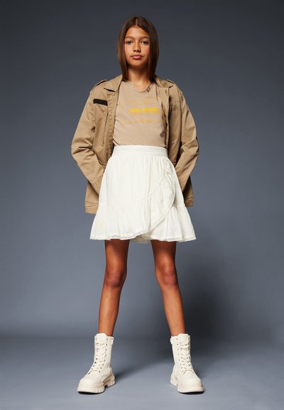 Veste beige, T-shirt marron clair avec texte jaune, jupe blanche à couches, et bottes crème hautes jusqu'aux genoux. Design simple et textures douces.