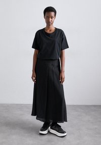 T-shirt nera a maniche corte e gonna maxi plissettata, entrambi in nero uniforme. L'outfit è abbinato a sneaker nere chunky con suola bianca.
