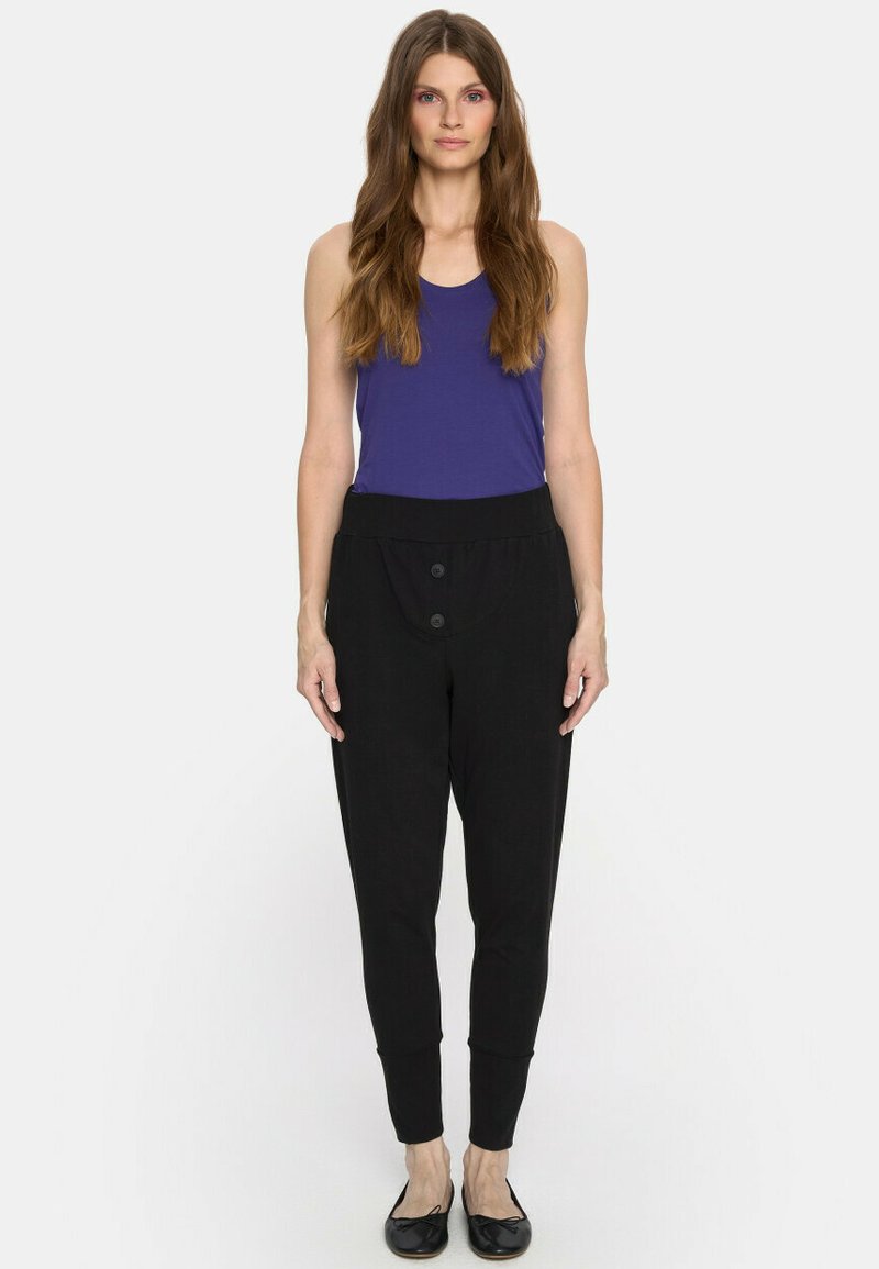 Kobieta stoi w bezrękawowej fioletowej bluzce, w luźnych czarnych spodniach z wysokim stanem z guzikami oraz czarnych baletkach, na białym tle.