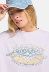 Camiseta de color púrpura claro con un gráfico circular azul y amarillo que presenta la palabra "BILLABONG". Modelo usando una gorra de mezclilla, sosteniendo el borde.