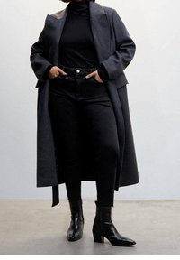 Abrigo largo negro de tela gruesa, superpuesto sobre un suéter negro de cuello alto y jeans negros de tiro alto; combinado con botas negras de tobillo.